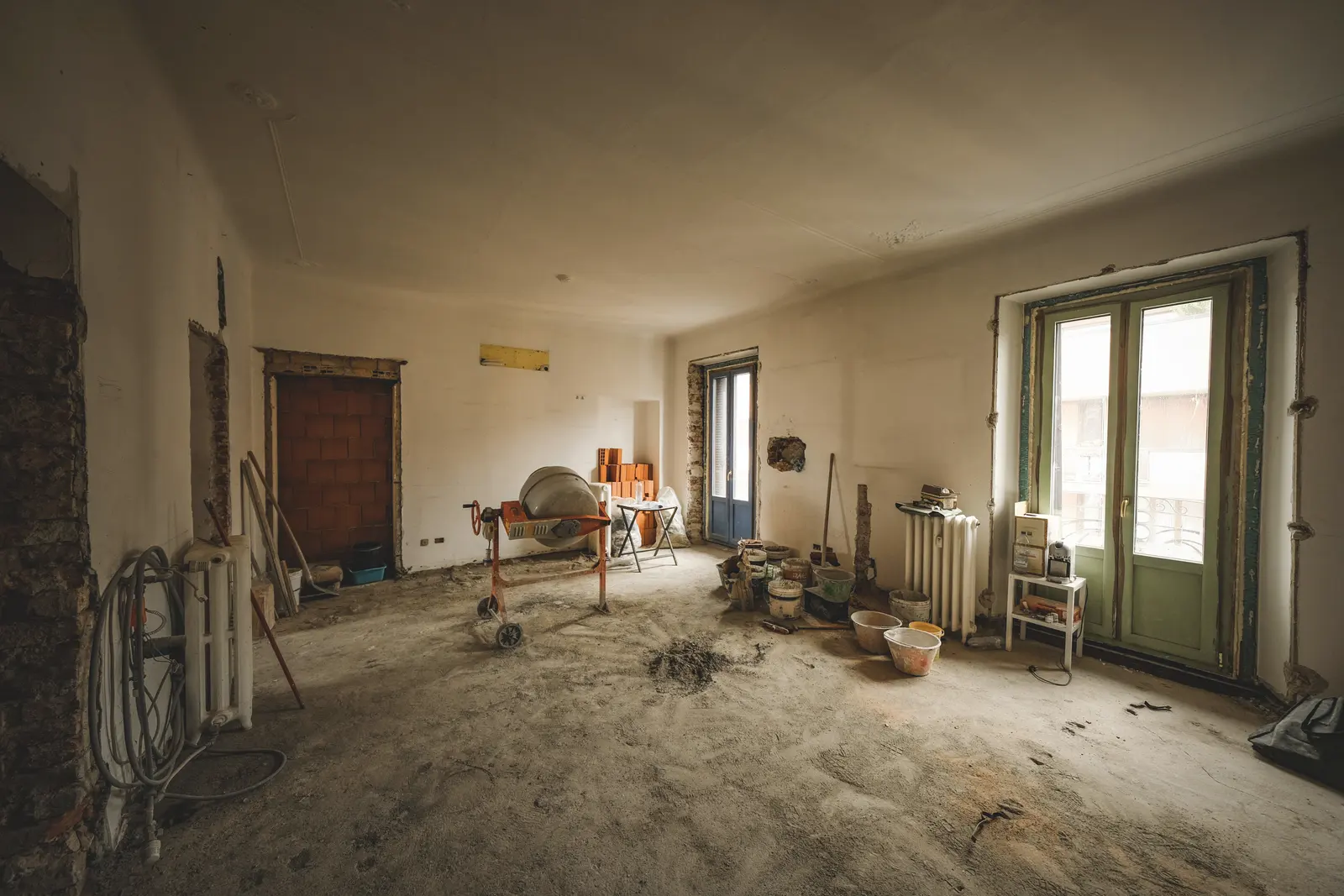 Une pièce avant travaux : sol brut, bétonnière, porte-fenêtre au fond.